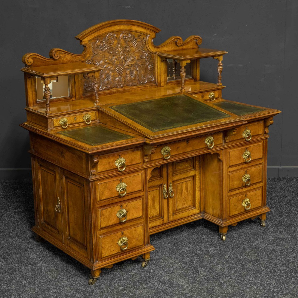 A late Victorian pollard oak desk featuring a central sloped green leather writing surface, carved backboard cresting, tiered shelves with mirrors, and brass-handled drawers.