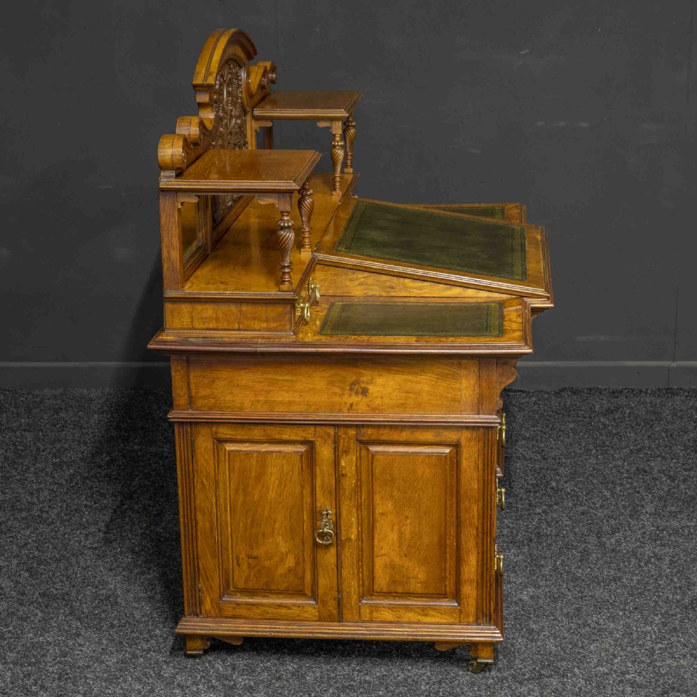 A late Victorian pollard oak desk featuring a central sloped green leather writing surface, carved backboard cresting, tiered shelves with mirrors, and brass-handled drawers. View IV