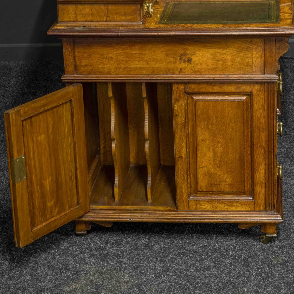 A late Victorian pollard oak desk featuring a central sloped green leather writing surface, carved backboard cresting, tiered shelves with mirrors, and brass-handled drawers. View V