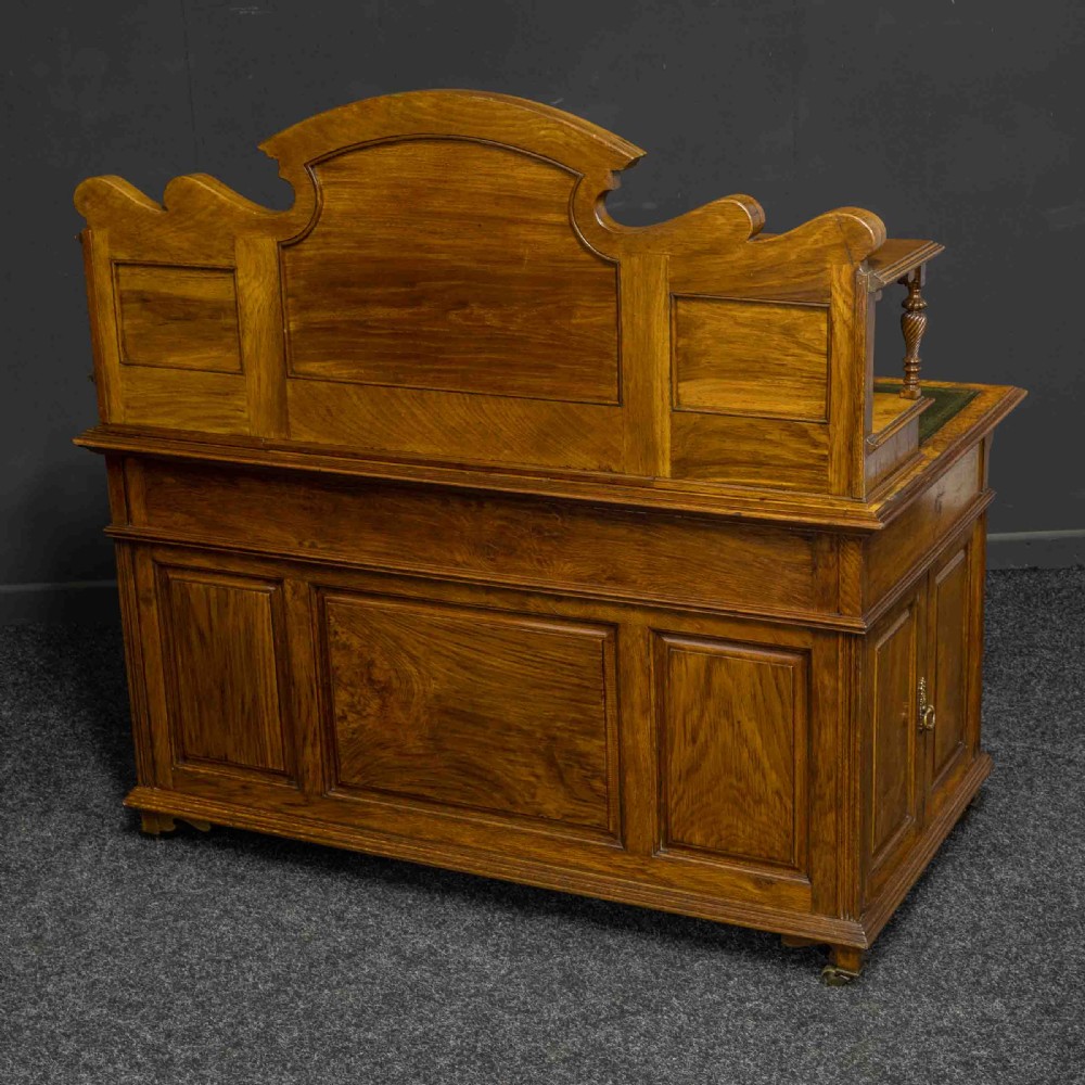 A late Victorian pollard oak desk featuring a central sloped green leather writing surface, carved backboard cresting, tiered shelves with mirrors, and brass-handled drawers. View VI