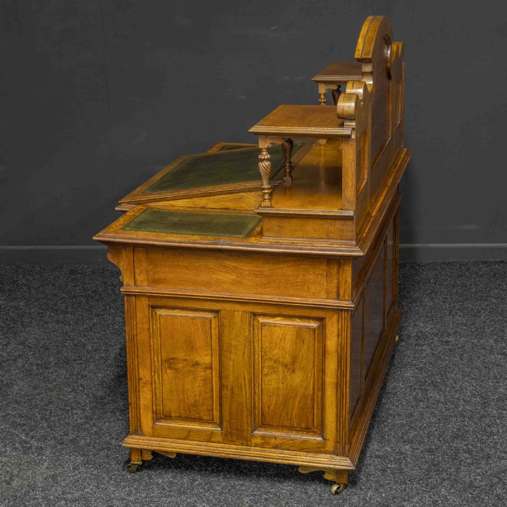 A late Victorian pollard oak desk featuring a central sloped green leather writing surface, carved backboard cresting, tiered shelves with mirrors, and brass-handled drawers. View VII