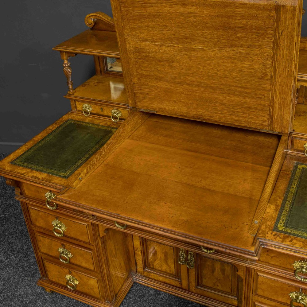 A late Victorian pollard oak desk featuring a central sloped green leather writing surface, carved backboard cresting, tiered shelves with mirrors, and brass-handled drawers. View XIII