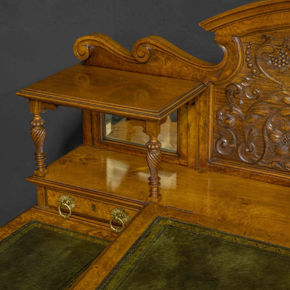 A late Victorian pollard oak desk featuring a central sloped green leather writing surface, carved backboard cresting, tiered shelves with mirrors, and brass-handled drawers. View XV
