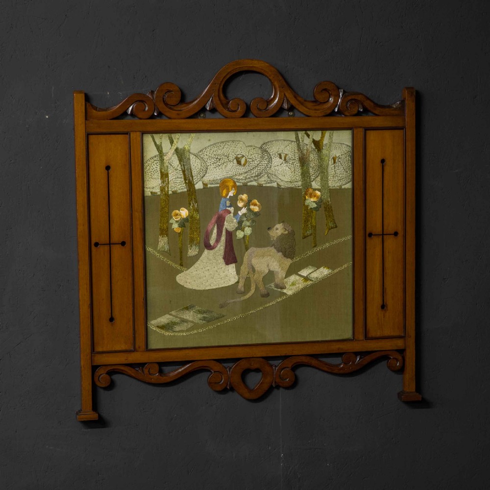 An Edwardian carved mahogany overmantel frame featuring a central embroidered panel depicting a figure with a lion, topped with scrollwork and an inverted heart cutout at the base.