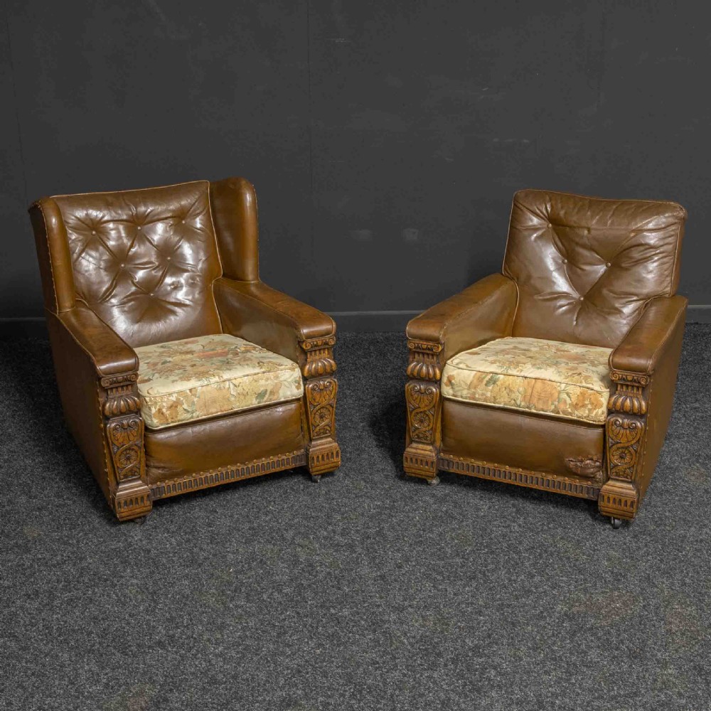 A pair of early 20th-century tan leather armchairs featuring buttoned backs, carved timber front pilasters, and floral patterned fabric seat cushions.