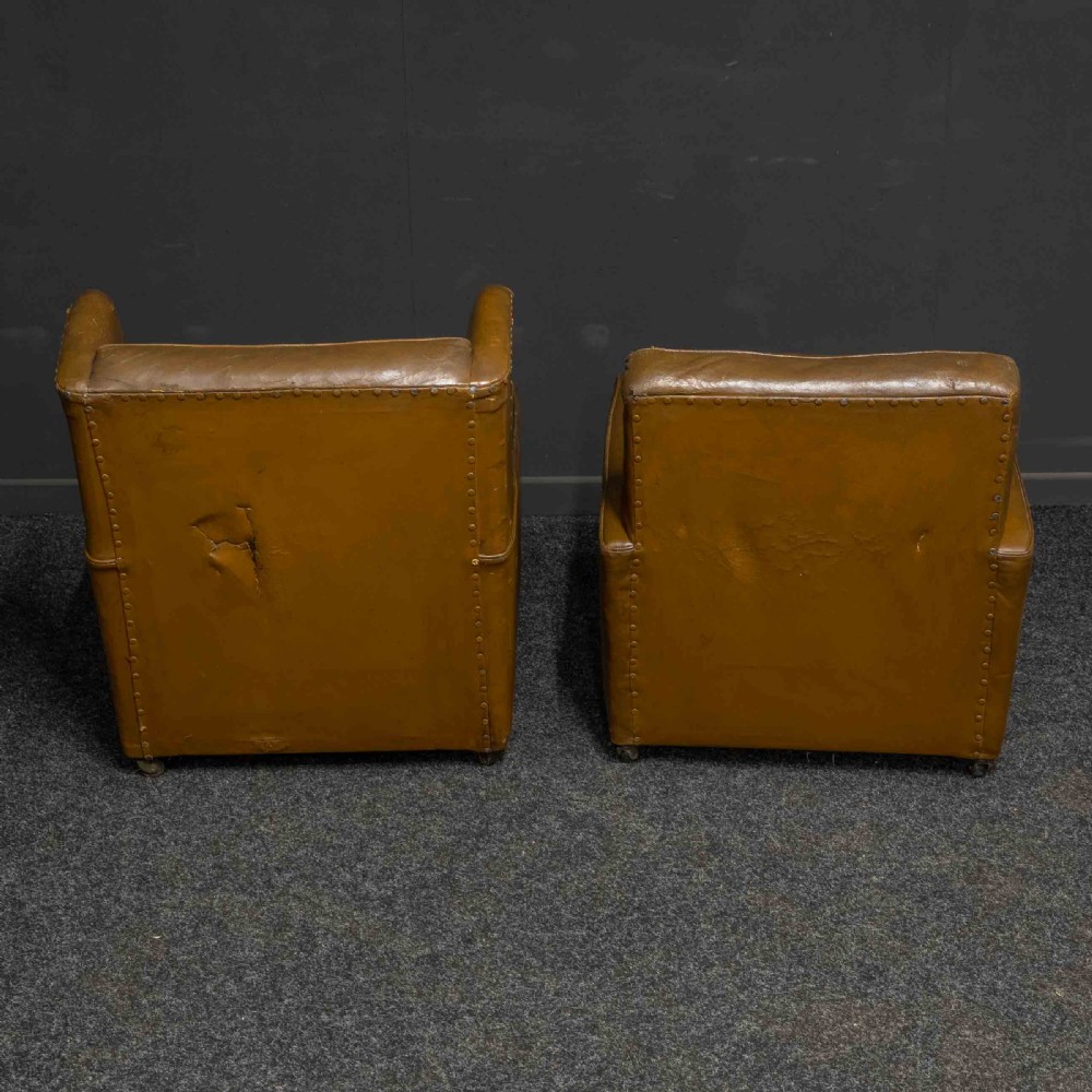 A pair of early 20th-century tan leather armchairs featuring buttoned backs, carved timber front pilasters, and floral patterned fabric seat cushions. View X