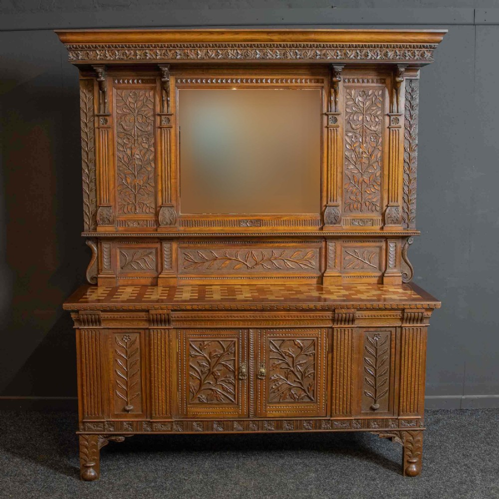 A carved oak Arts and Crafts dresser featuring a mirror back upper section, carved leaf panels, checkerboard inlay counter, and lower cupboards.