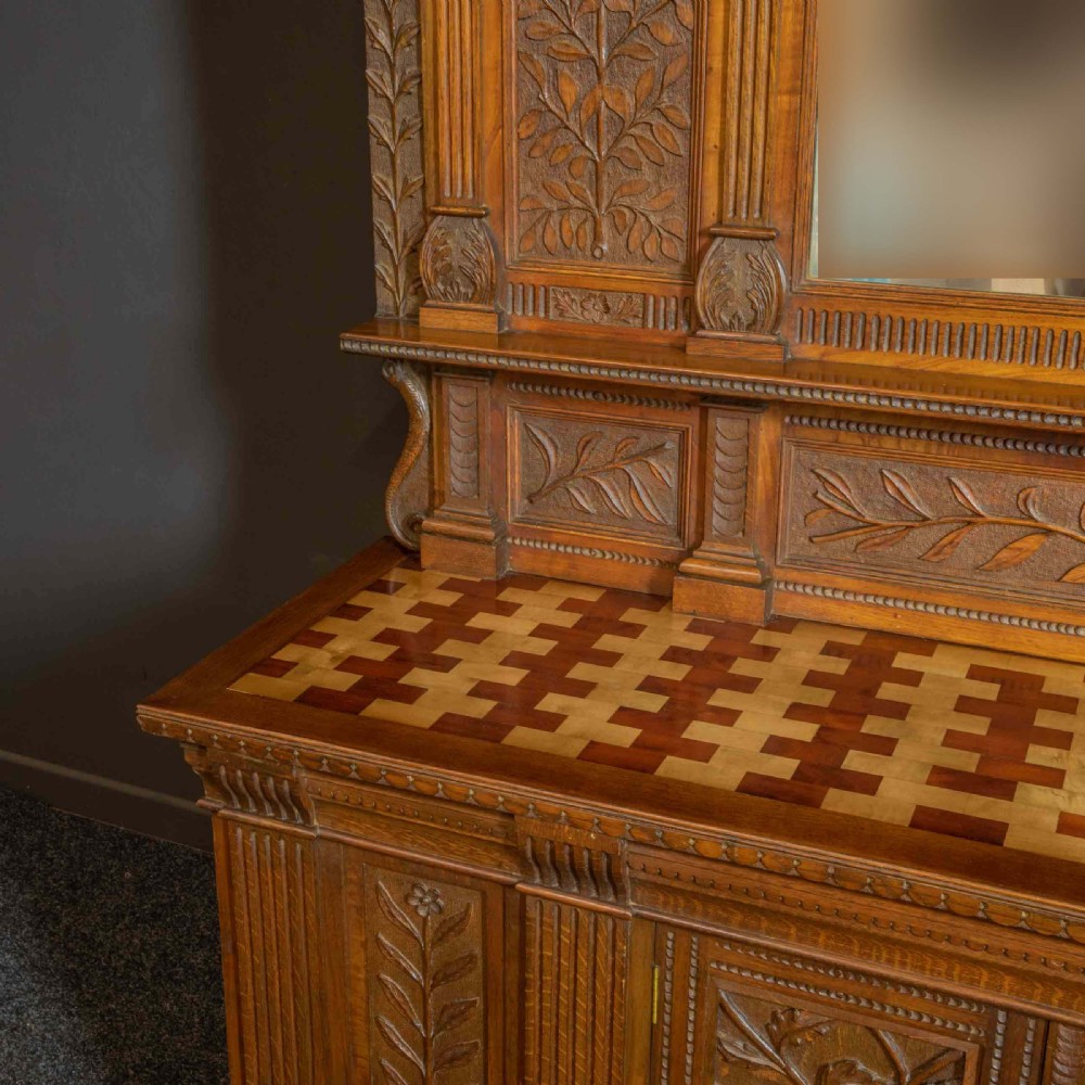 A carved oak Arts and Crafts dresser featuring a mirror back upper section, carved leaf panels, checkerboard inlay counter, and lower cupboards. View II
