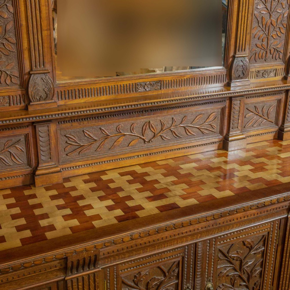 A carved oak Arts and Crafts dresser featuring a mirror back upper section, carved leaf panels, checkerboard inlay counter, and lower cupboards. View III