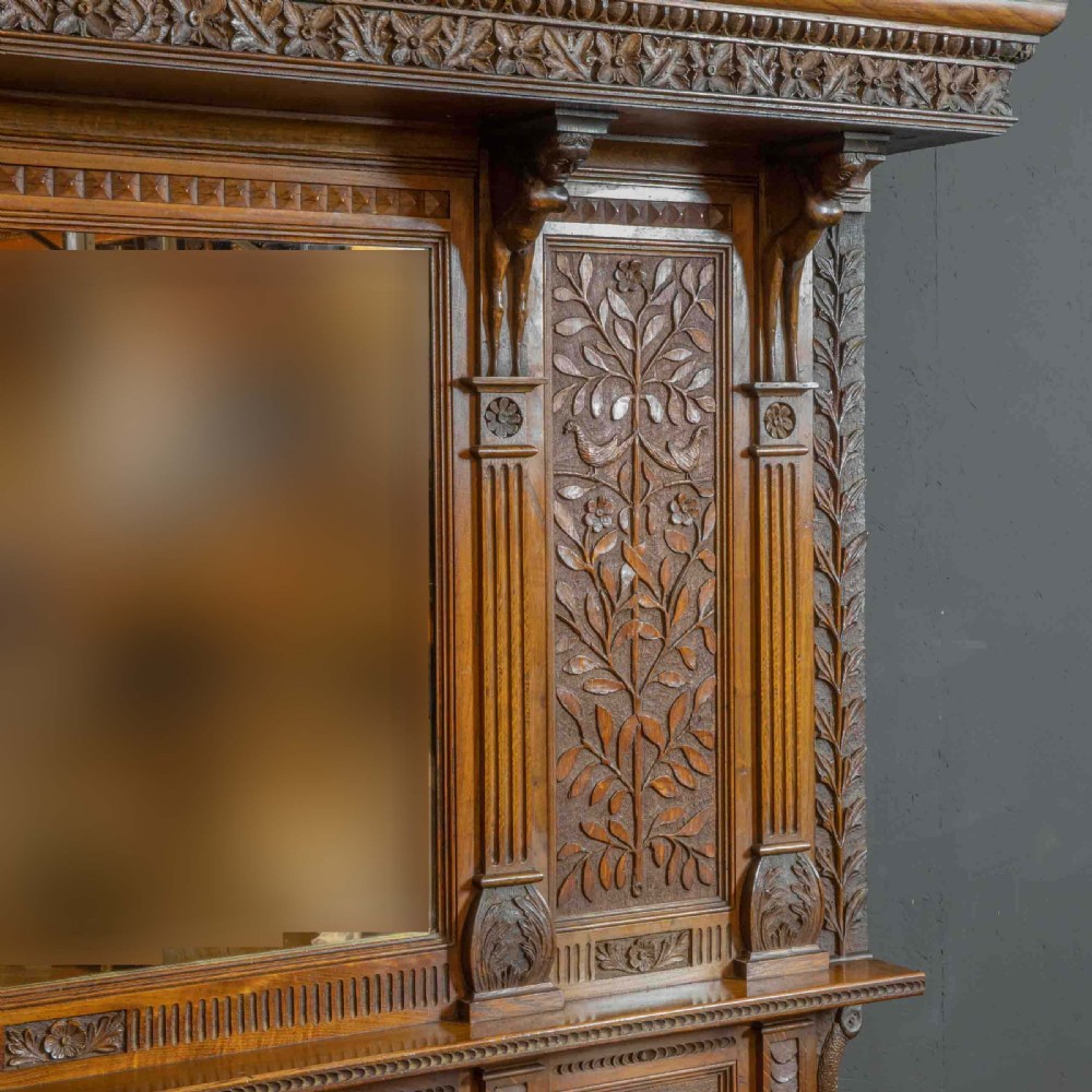 A carved oak Arts and Crafts dresser featuring a mirror back upper section, carved leaf panels, checkerboard inlay counter, and lower cupboards. View V