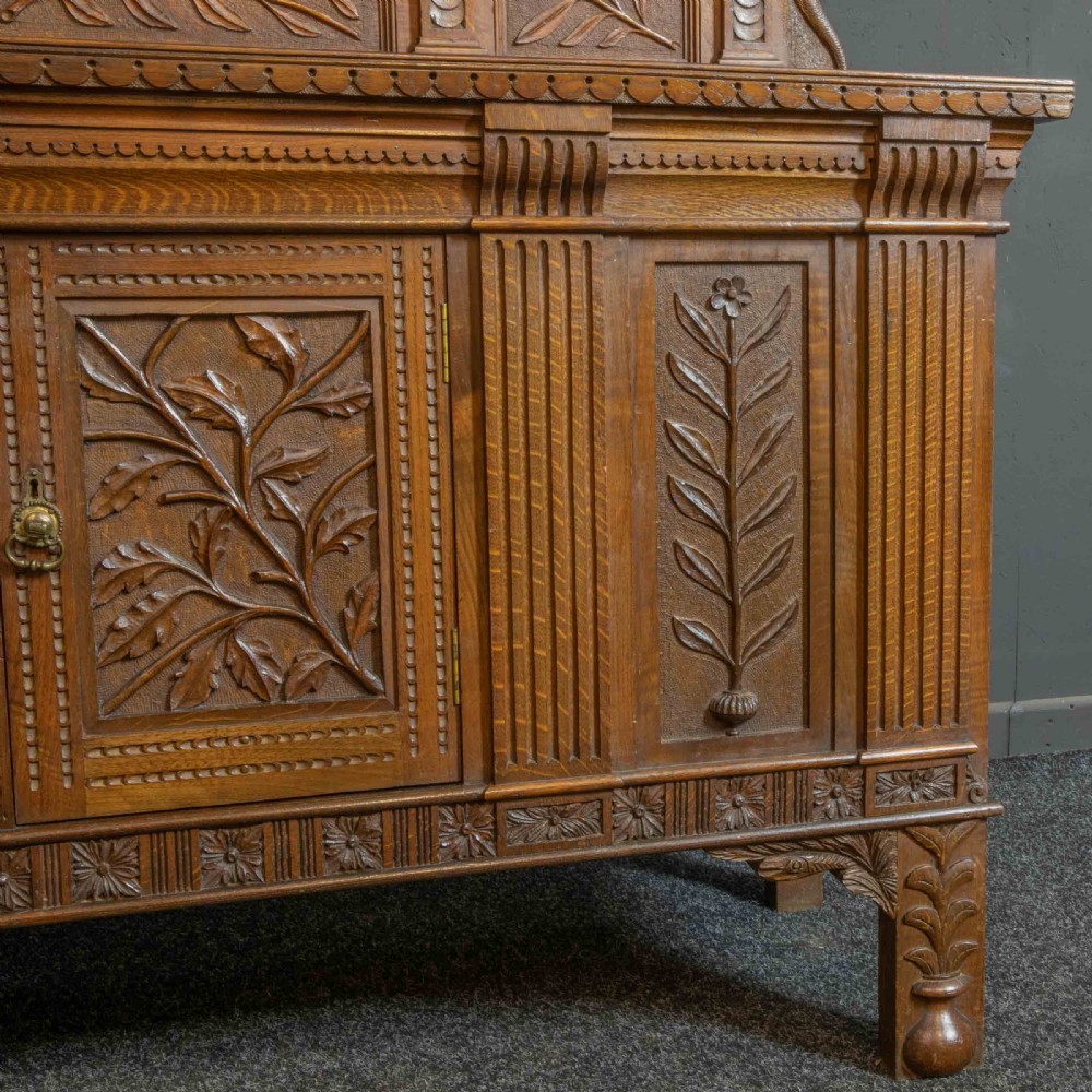 A carved oak Arts and Crafts dresser featuring a mirror back upper section, carved leaf panels, checkerboard inlay counter, and lower cupboards. View VIII