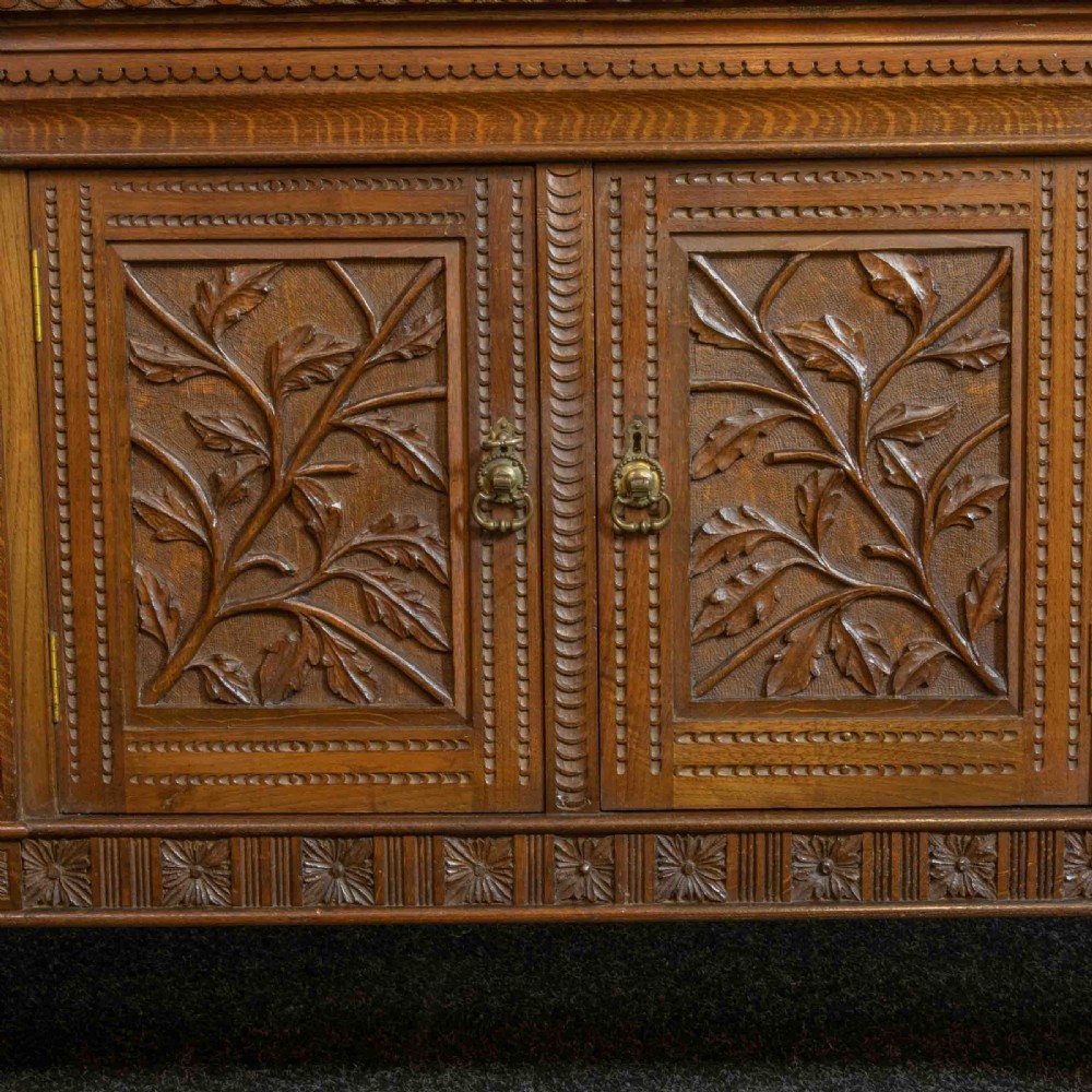 A carved oak Arts and Crafts dresser featuring a mirror back upper section, carved leaf panels, checkerboard inlay counter, and lower cupboards. View IX