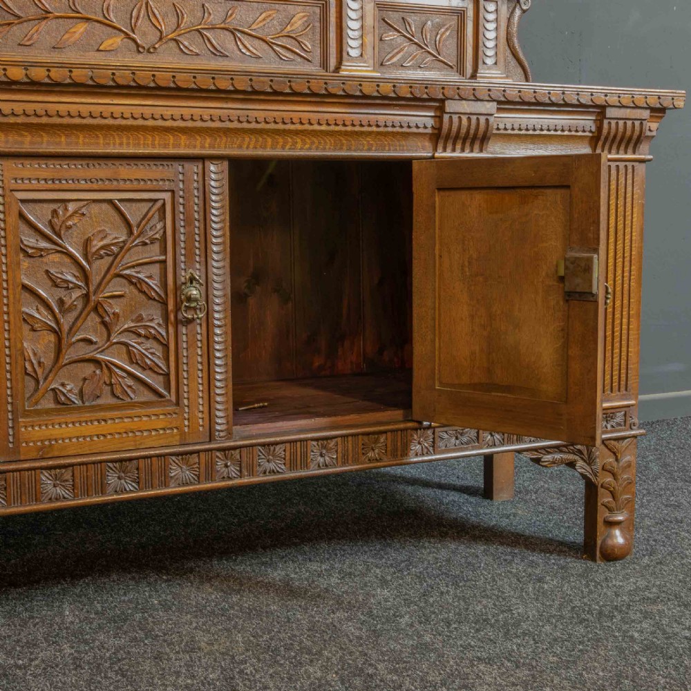 A carved oak Arts and Crafts dresser featuring a mirror back upper section, carved leaf panels, checkerboard inlay counter, and lower cupboards. View X