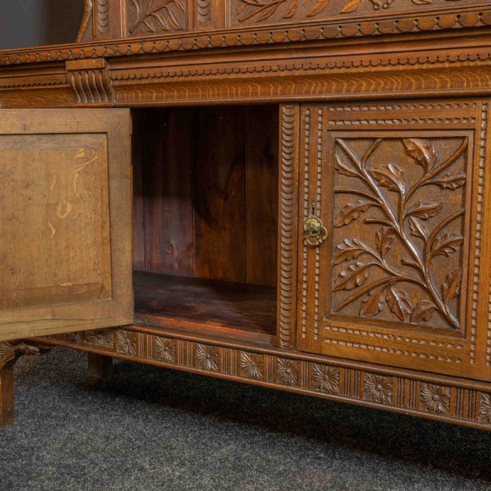 A carved oak Arts and Crafts dresser featuring a mirror back upper section, carved leaf panels, checkerboard inlay counter, and lower cupboards. View XI