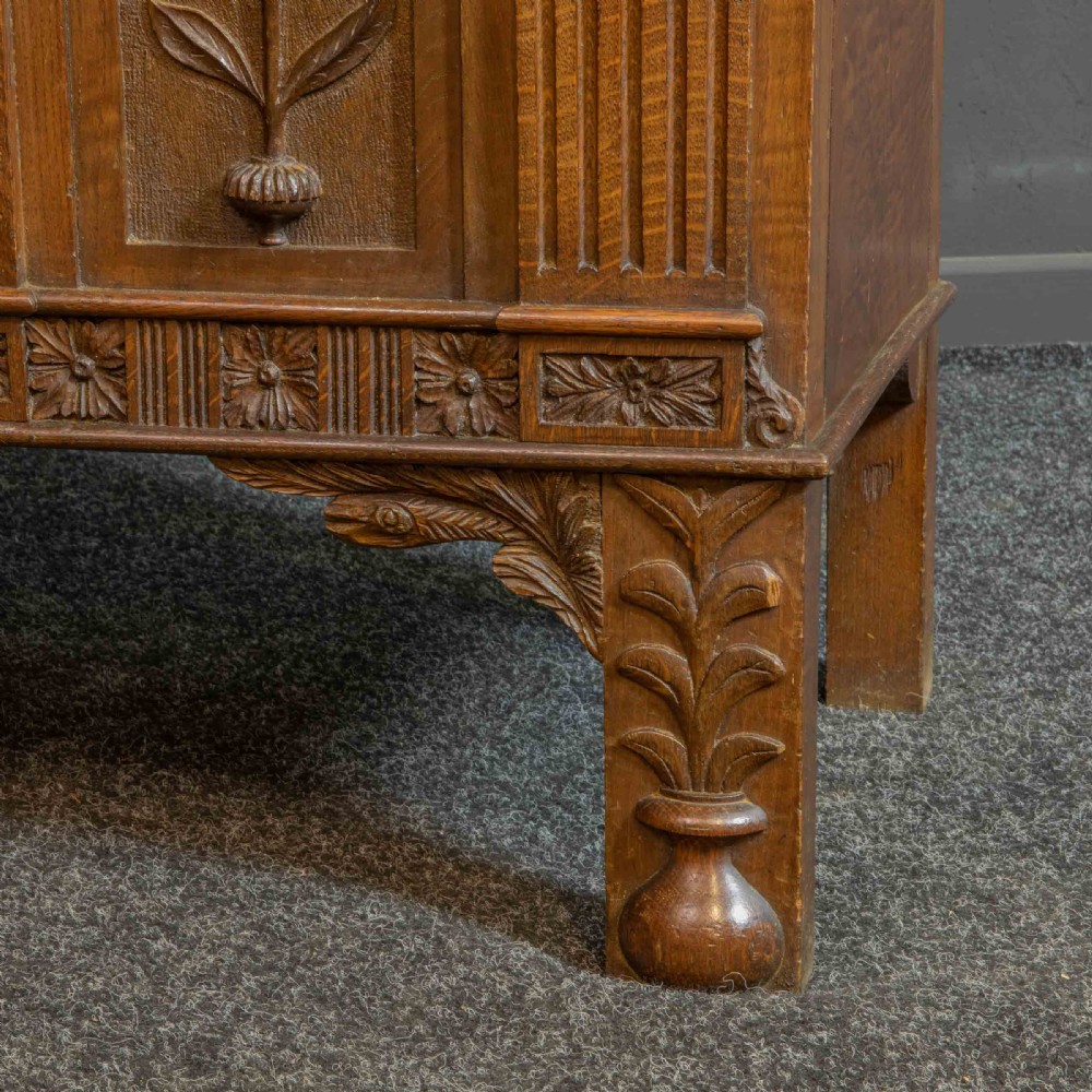 A carved oak Arts and Crafts dresser featuring a mirror back upper section, carved leaf panels, checkerboard inlay counter, and lower cupboards. View XII