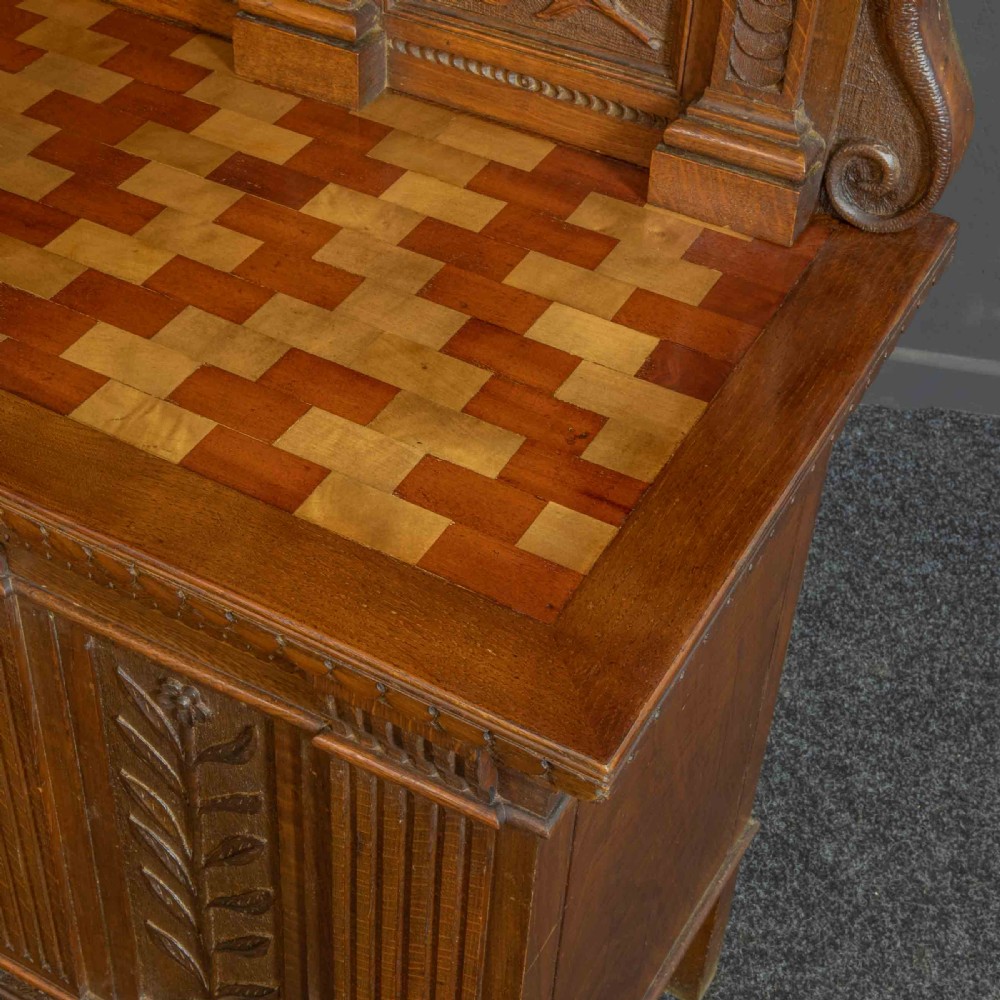 A carved oak Arts and Crafts dresser featuring a mirror back upper section, carved leaf panels, checkerboard inlay counter, and lower cupboards. View XIII