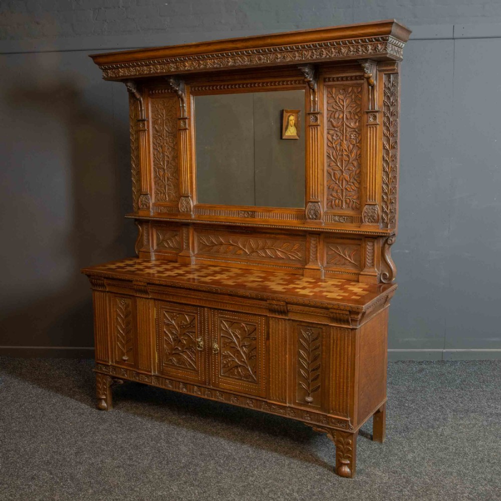 A carved oak Arts and Crafts dresser featuring a mirror back upper section, carved leaf panels, checkerboard inlay counter, and lower cupboards. View XV