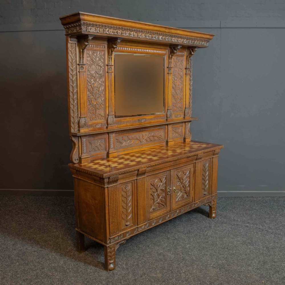 A carved oak Arts and Crafts dresser featuring a mirror back upper section, carved leaf panels, checkerboard inlay counter, and lower cupboards. View XVI