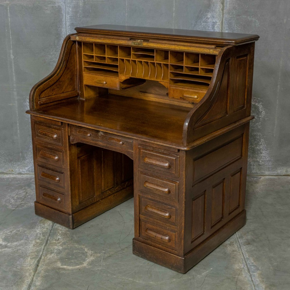 An Edwardian dark oak roll top desk featuring an open slatted tambour hood, fitted interior pigeonhole compartments, and twin pedestals with carved wooden drawer handles.