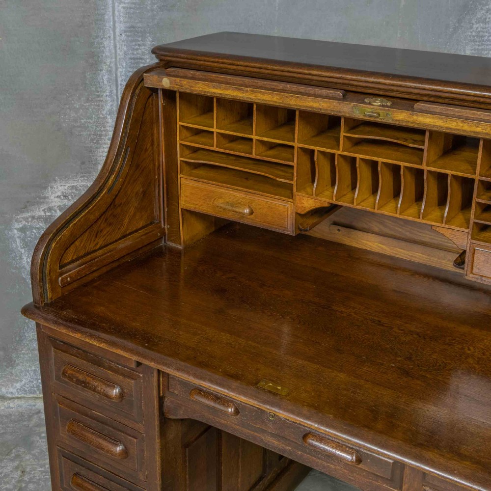 An Edwardian dark oak roll top desk featuring an open slatted tambour hood, fitted interior pigeonhole compartments, and twin pedestals with carved wooden drawer handles. View III