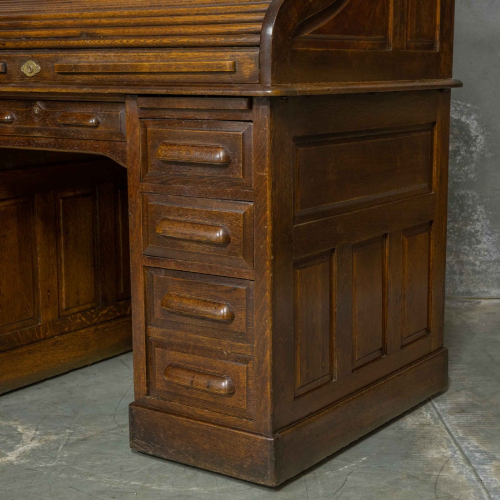 An Edwardian dark oak roll top desk featuring an open slatted tambour hood, fitted interior pigeonhole compartments, and twin pedestals with carved wooden drawer handles. View XII