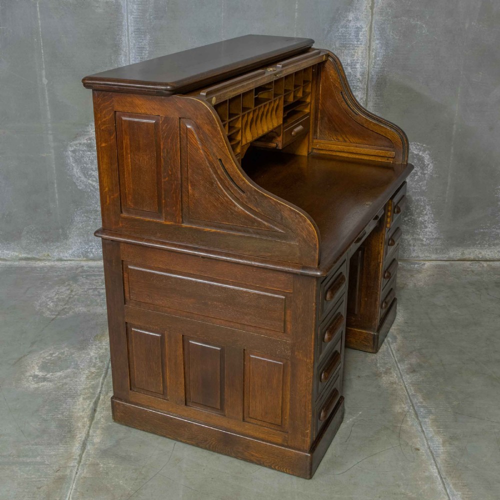 An Edwardian dark oak roll top desk featuring an open slatted tambour hood, fitted interior pigeonhole compartments, and twin pedestals with carved wooden drawer handles. View XV