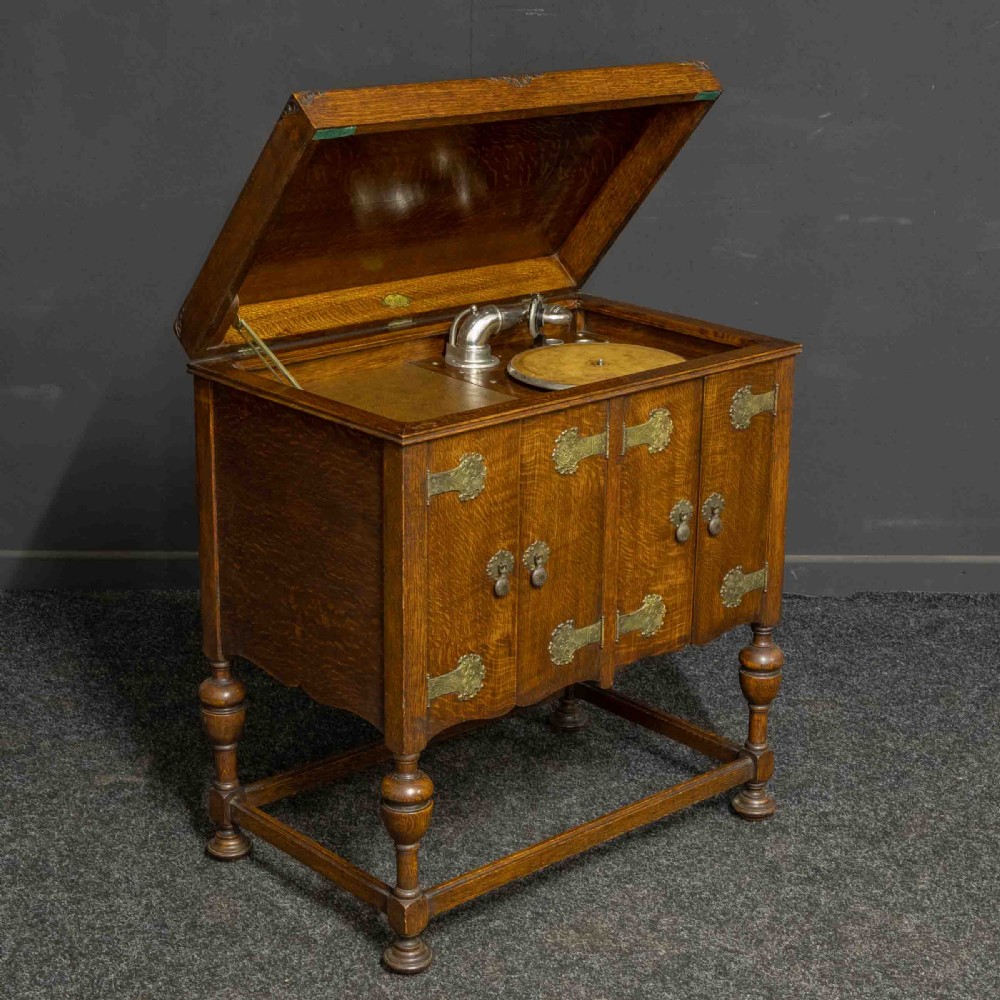 An early 20th-century medium oak wind-up cabinet gramophone with brass strap hinges, drop handles, turned legs, and an open top lid showing the turntable mechanism.