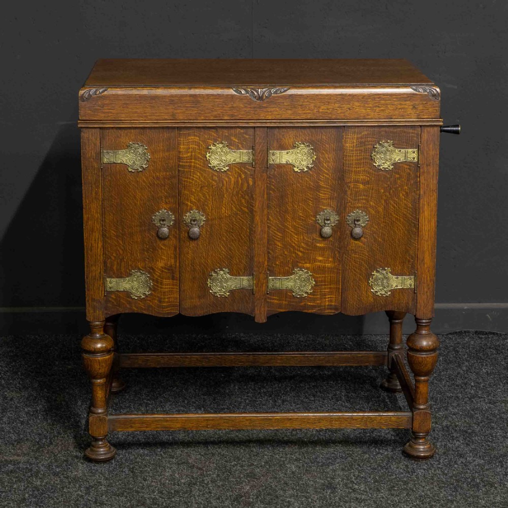 An early 20th-century medium oak wind-up cabinet gramophone with brass strap hinges, drop handles, turned legs, and an open top lid showing the turntable mechanism. View IV