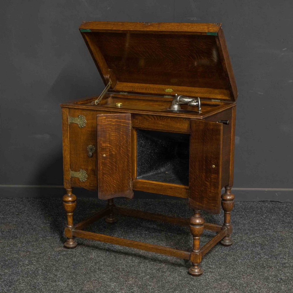 An early 20th-century medium oak wind-up cabinet gramophone with brass strap hinges, drop handles, turned legs, and an open top lid showing the turntable mechanism. View V