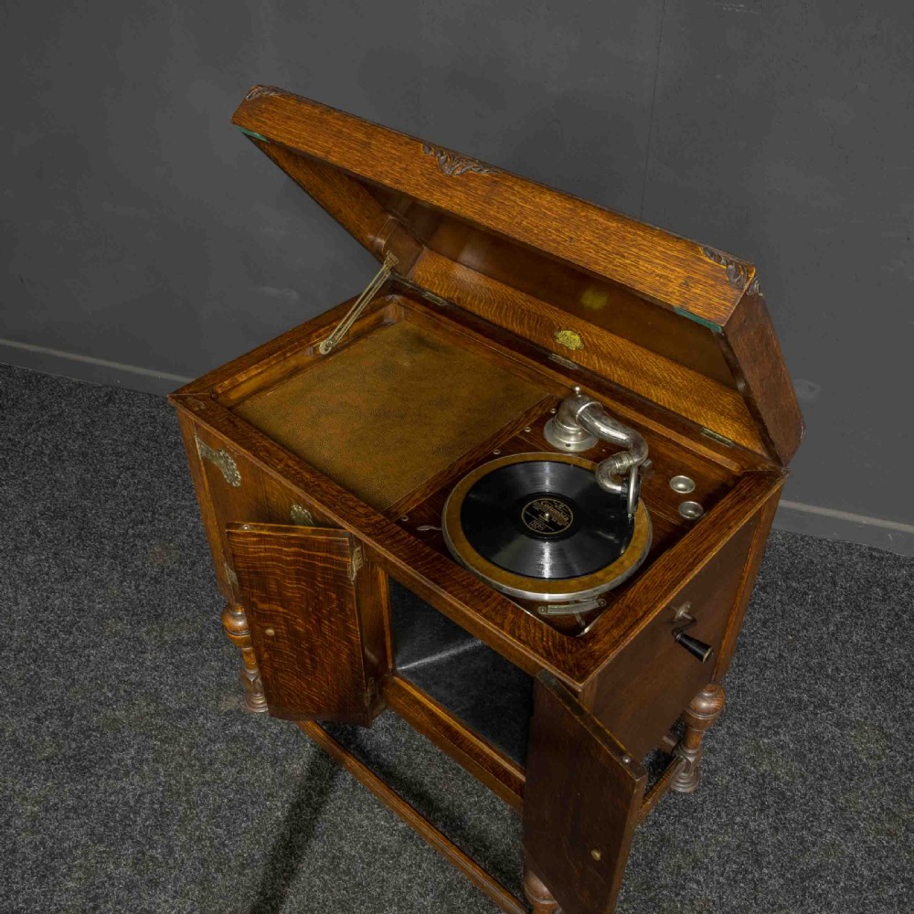 An early 20th-century medium oak wind-up cabinet gramophone with brass strap hinges, drop handles, turned legs, and an open top lid showing the turntable mechanism. View VII