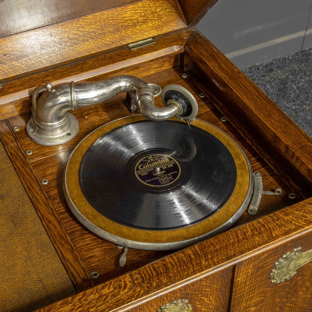 An early 20th-century medium oak wind-up cabinet gramophone with brass strap hinges, drop handles, turned legs, and an open top lid showing the turntable mechanism. View X