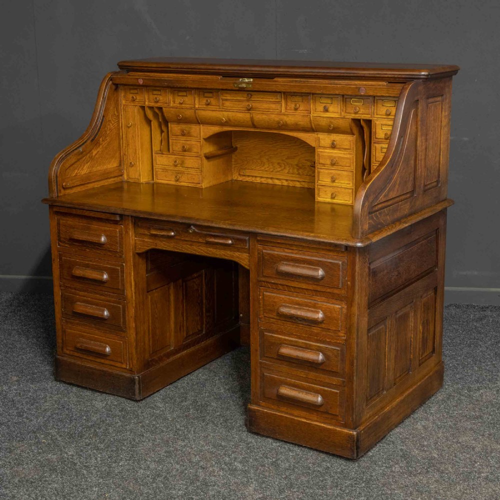 An Edwardian oak roll top desk featuring an S-curve slatted shutter opening to a fitted interior with small drawers and pigeonholes above twin pedestal drawers.
