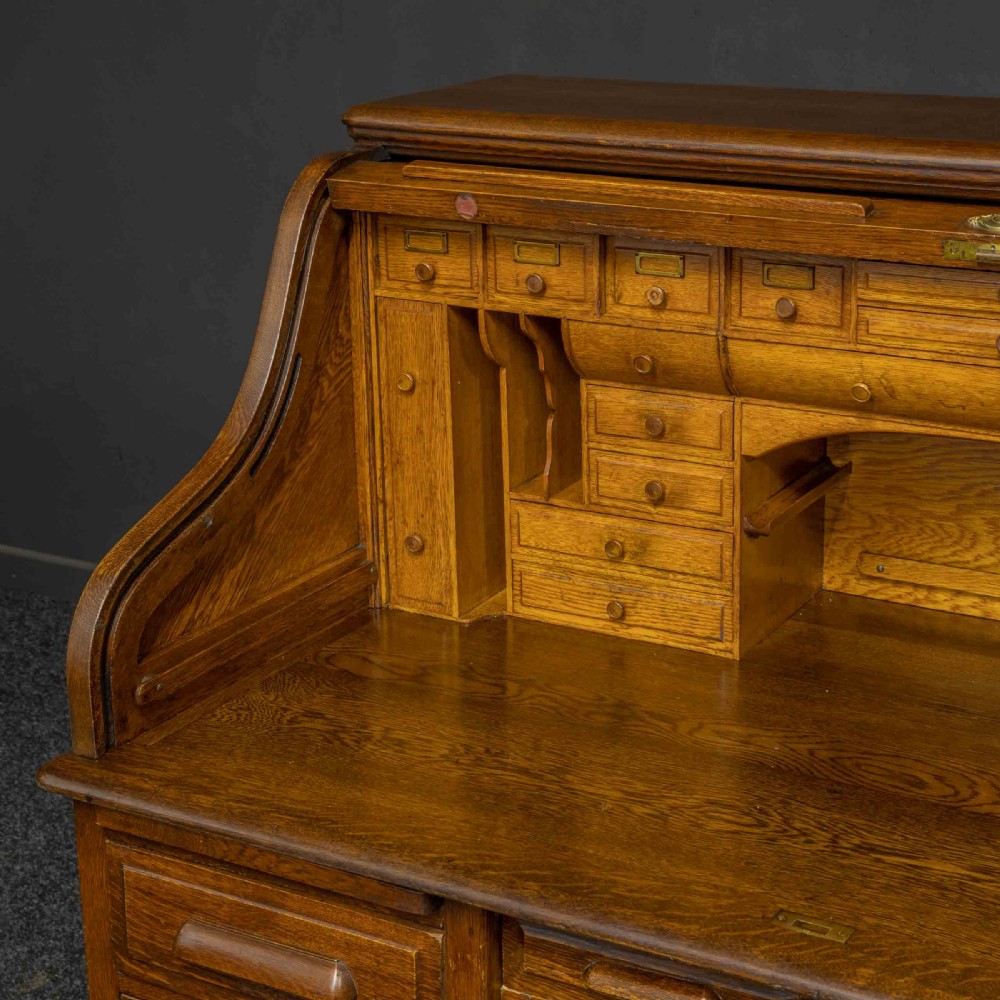An Edwardian oak roll top desk featuring an S-curve slatted shutter opening to a fitted interior with small drawers and pigeonholes above twin pedestal drawers. View IV