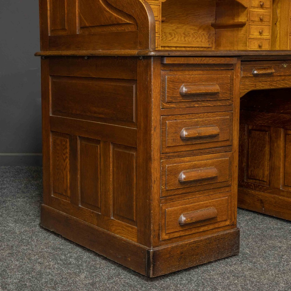 An Edwardian oak roll top desk featuring an S-curve slatted shutter opening to a fitted interior with small drawers and pigeonholes above twin pedestal drawers. View IX