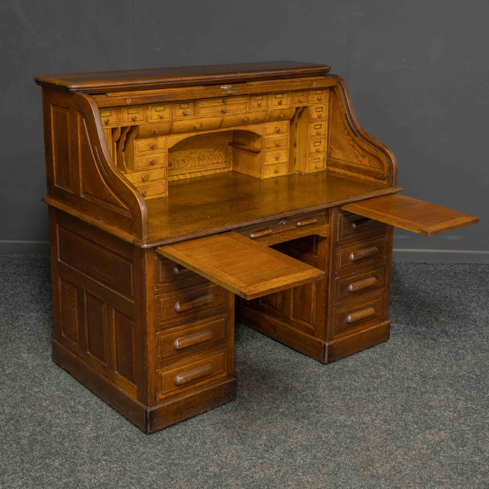 An Edwardian oak roll top desk featuring an S-curve slatted shutter opening to a fitted interior with small drawers and pigeonholes above twin pedestal drawers. View X