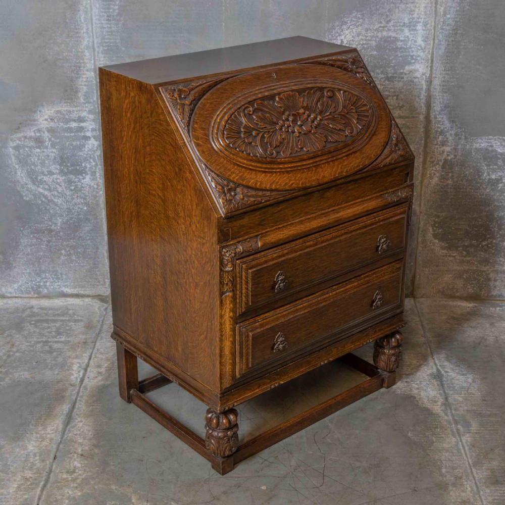 An early 20th-century dark oak writing bureau with a carved oval floral motif on the sloped flap above two drawers.
