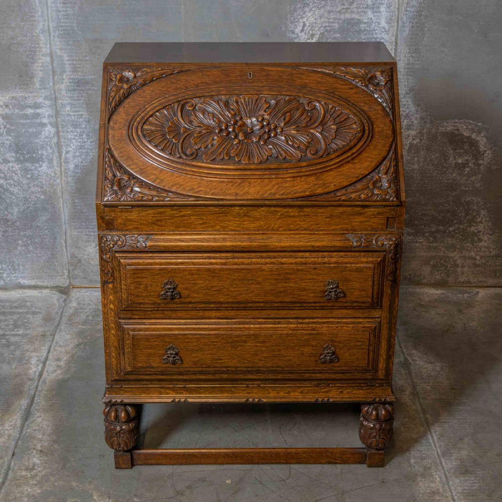 An early 20th-century dark oak writing bureau with a carved oval floral motif on the sloped flap above two drawers. View IX