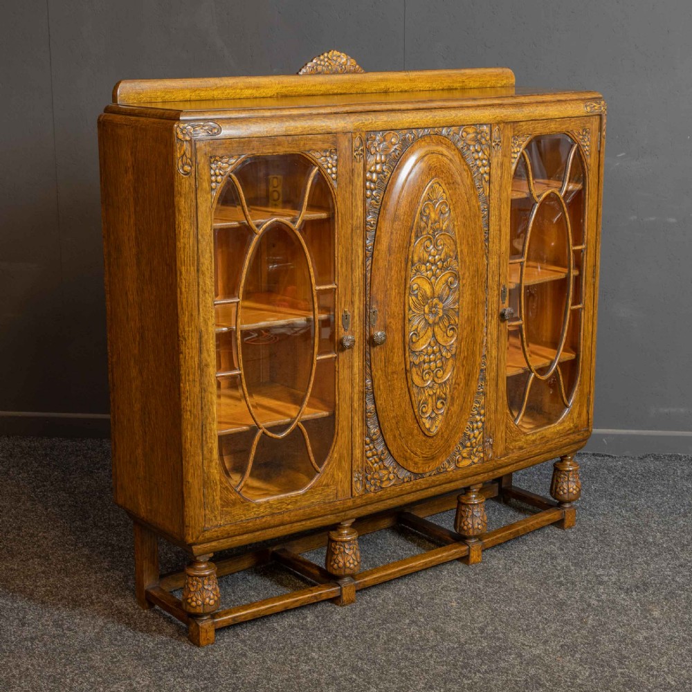An early 20th-century carved oak display cabinet with a central carved floral panel door flanked by two oval-pattern glazed doors set on turned legs with stretchers.