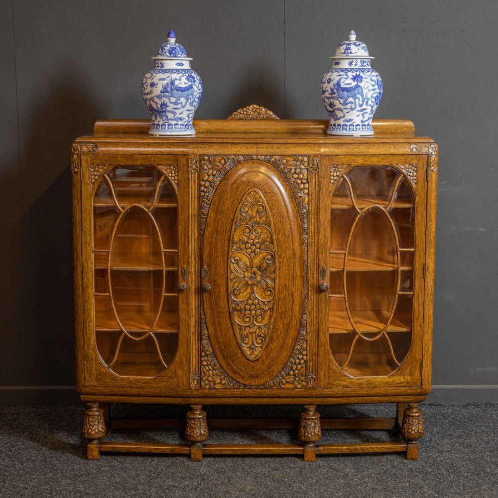 An early 20th-century carved oak display cabinet with a central carved floral panel door flanked by two oval-pattern glazed doors set on turned legs with stretchers. View II