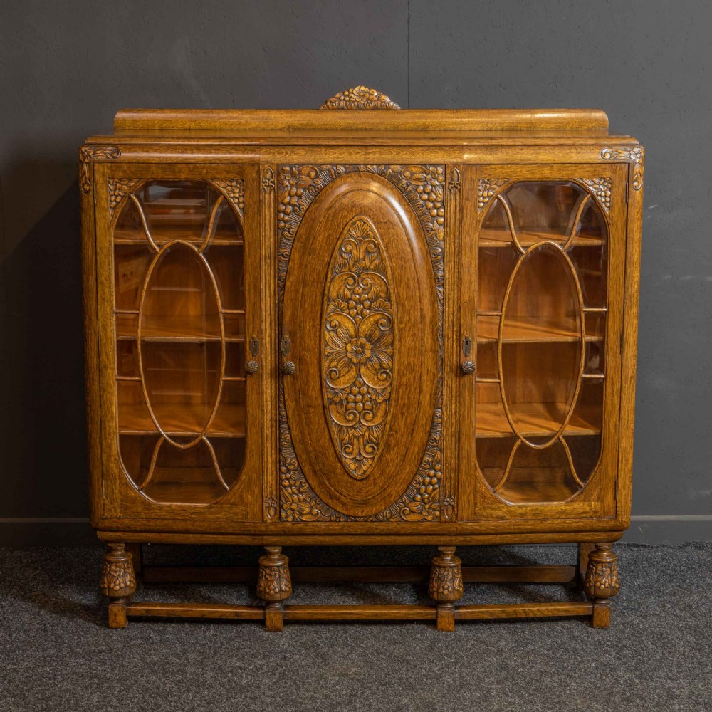 An early 20th-century carved oak display cabinet with a central carved floral panel door flanked by two oval-pattern glazed doors set on turned legs with stretchers. View III