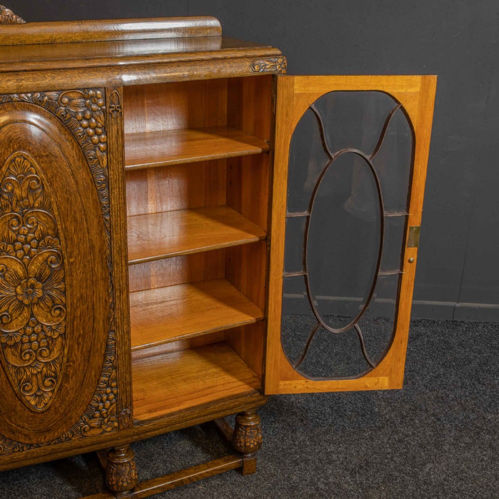 An early 20th-century carved oak display cabinet with a central carved floral panel door flanked by two oval-pattern glazed doors set on turned legs with stretchers. View VI