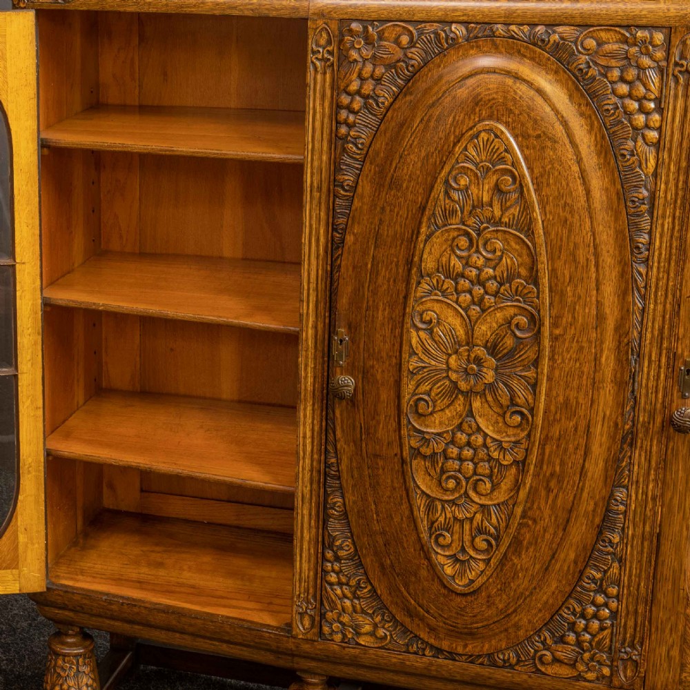An early 20th-century carved oak display cabinet with a central carved floral panel door flanked by two oval-pattern glazed doors set on turned legs with stretchers. View VII