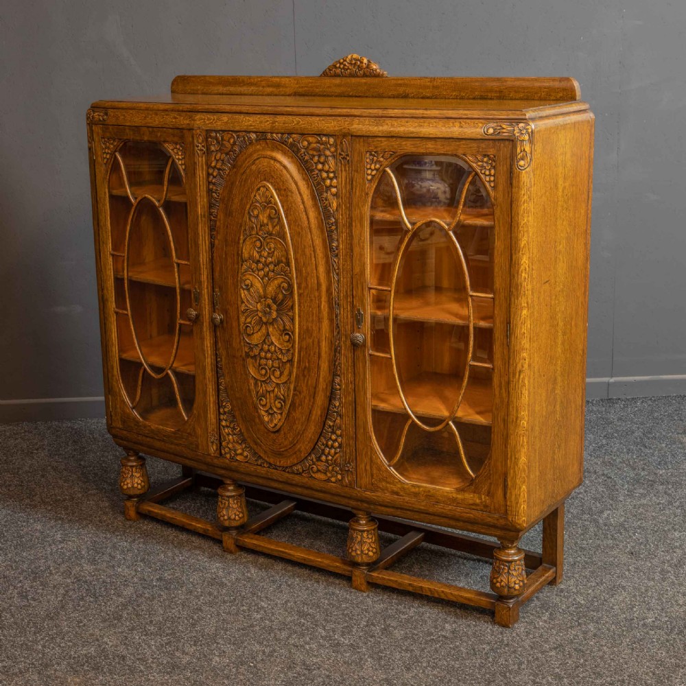 An early 20th-century carved oak display cabinet with a central carved floral panel door flanked by two oval-pattern glazed doors set on turned legs with stretchers. View VIII
