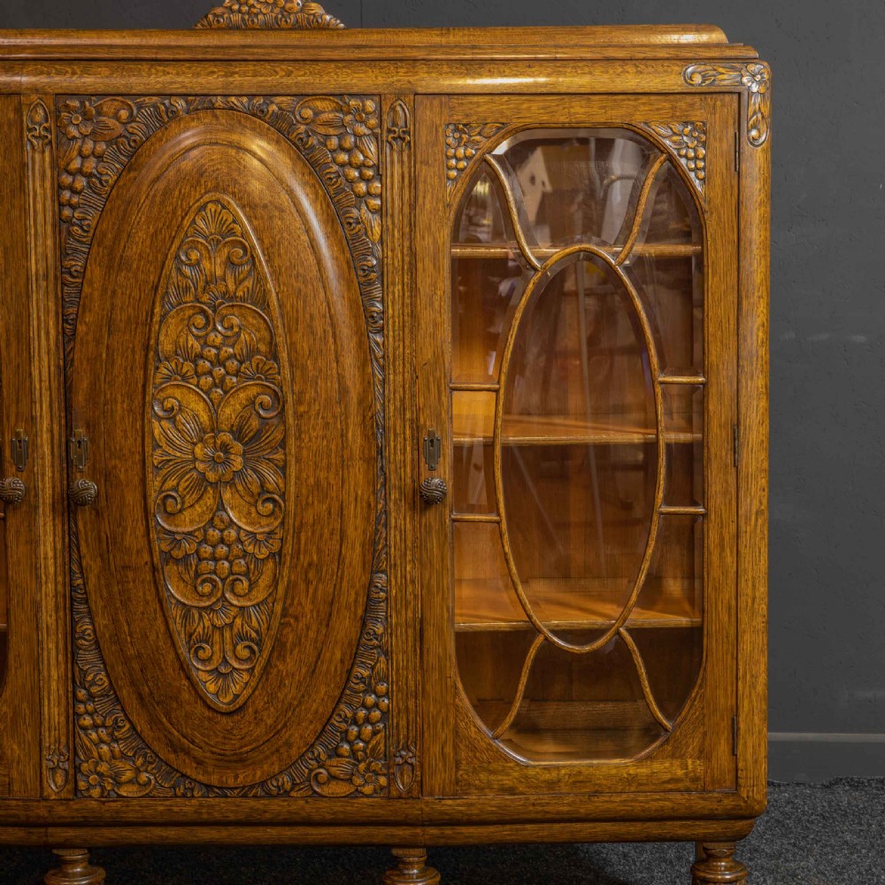An early 20th-century carved oak display cabinet with a central carved floral panel door flanked by two oval-pattern glazed doors set on turned legs with stretchers. View XI