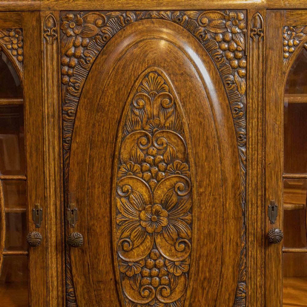 An early 20th-century carved oak display cabinet with a central carved floral panel door flanked by two oval-pattern glazed doors set on turned legs with stretchers. View XII