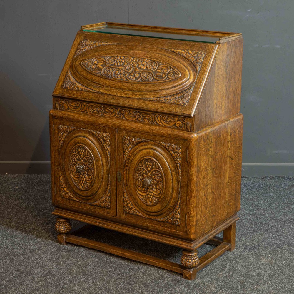 A medium brown carved oak writing bureau with a slant front lid above a double door cupboard, detailed with carved oval foliate panels and turned feet.