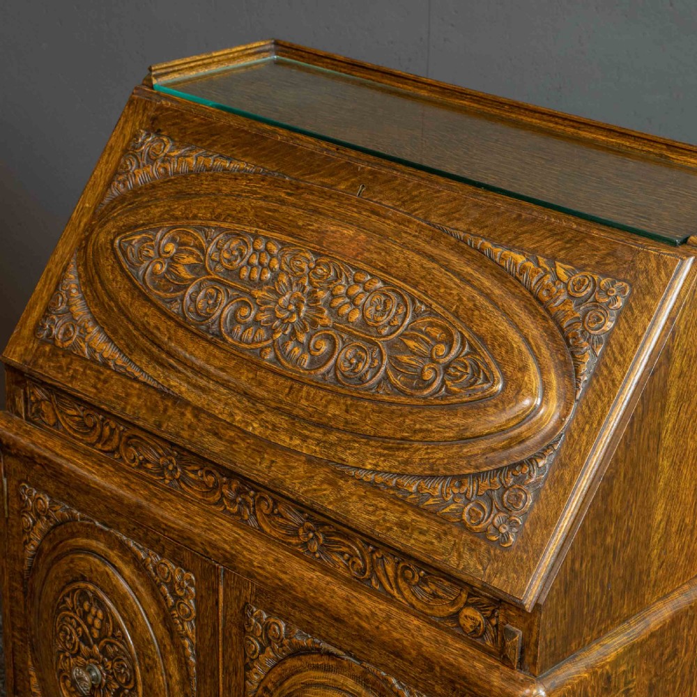 A medium brown carved oak writing bureau with a slant front lid above a double door cupboard, detailed with carved oval foliate panels and turned feet. View IV