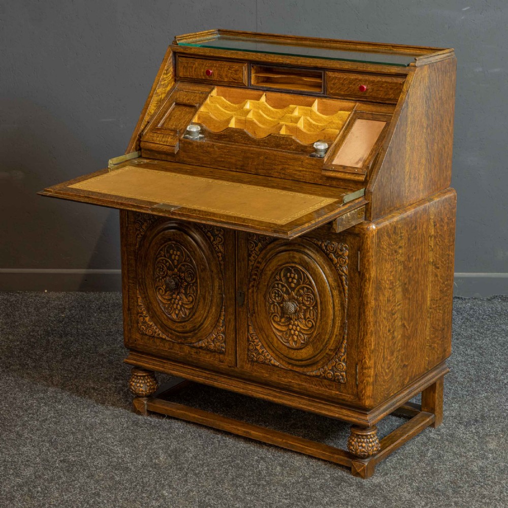 A medium brown carved oak writing bureau with a slant front lid above a double door cupboard, detailed with carved oval foliate panels and turned feet. View VI