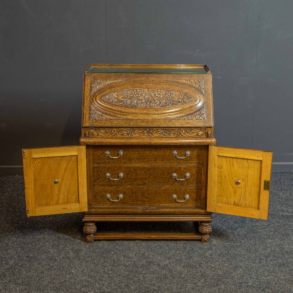 A medium brown carved oak writing bureau with a slant front lid above a double door cupboard, detailed with carved oval foliate panels and turned feet. View IX