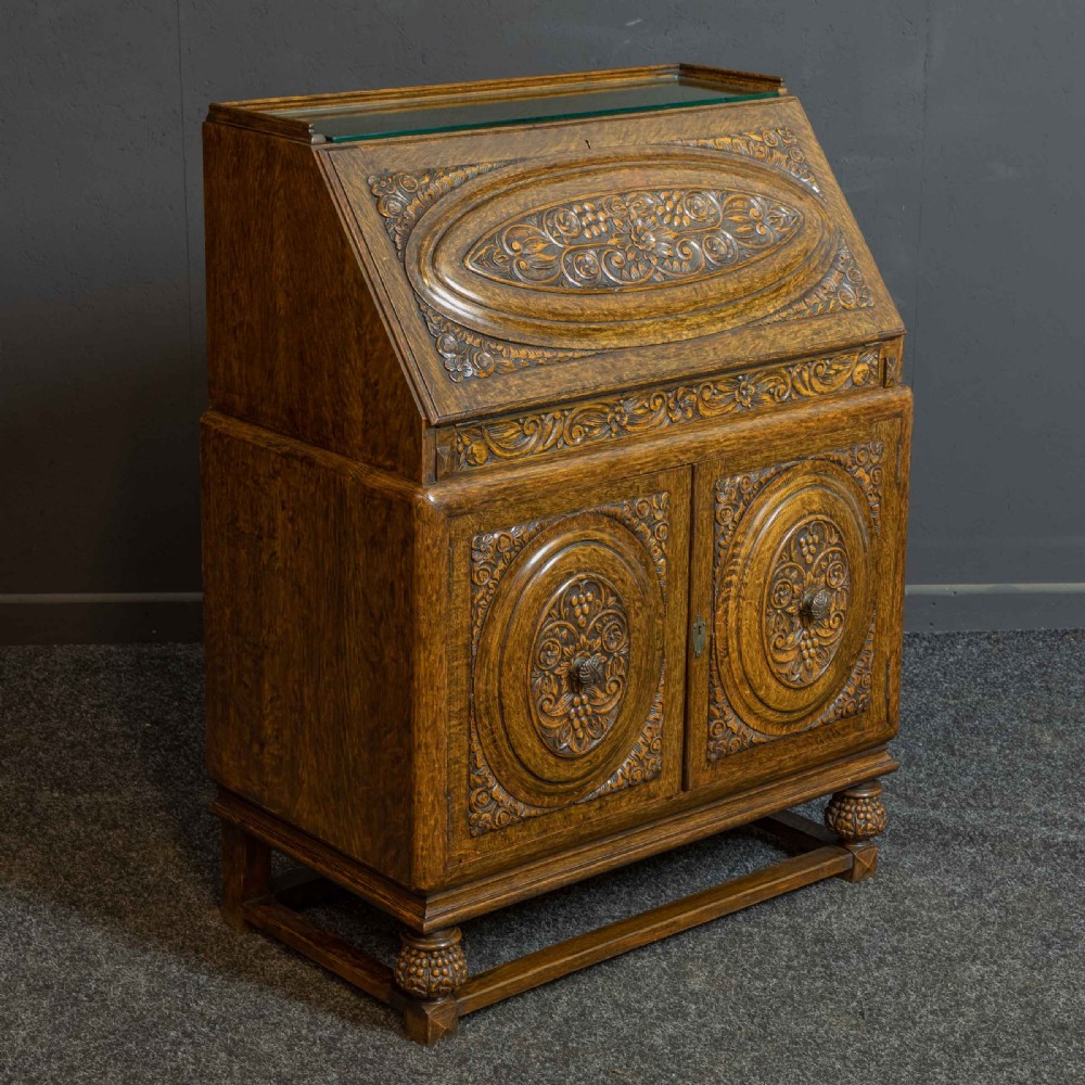 A medium brown carved oak writing bureau with a slant front lid above a double door cupboard, detailed with carved oval foliate panels and turned feet. View XIV